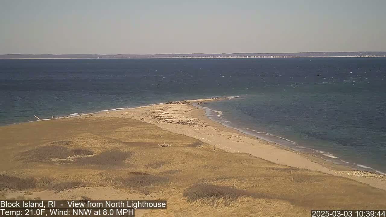 A sandy, narrow spit of land juts out into a calm, dark-blue ocean under a clear sky.