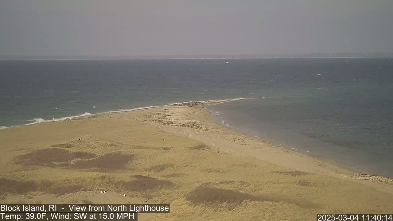 A sandy, grassy point of land juts into a calm, gray-blue ocean under a hazy sky, with a 15 mph southwest wind.