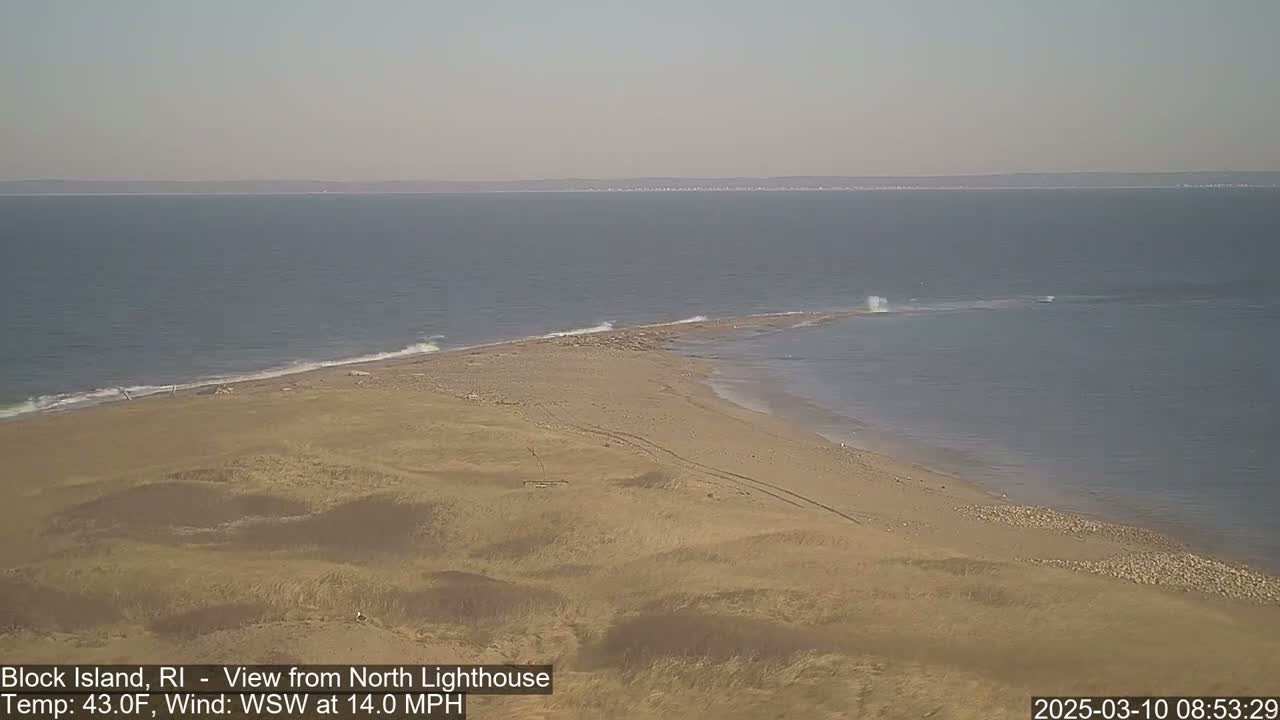 A sandy, dune-covered point of land juts into a calm ocean under a hazy sky, with a moderate wind blowing from the west-southwest.