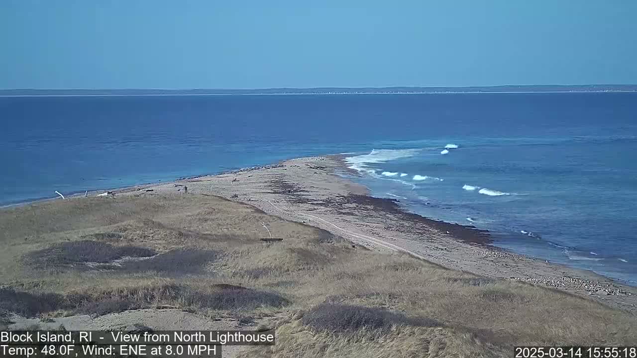 A sandy, grassy coastal point juts into a blue ocean with small waves under a clear sky.