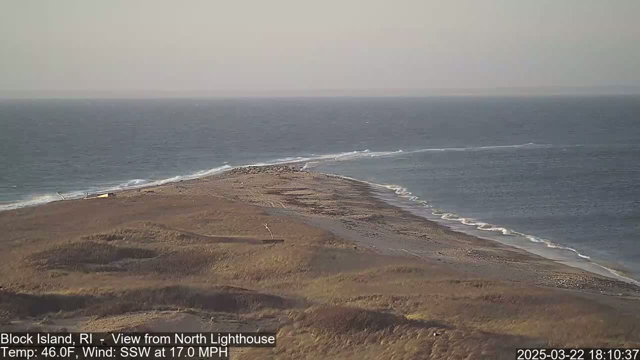 A sandy, grassy point of land juts into a wavy ocean under a mostly clear sky.