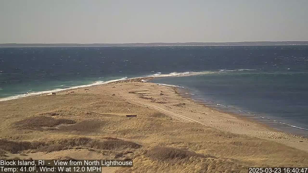 A sandy, grassy shoreline with a narrow isthmus extending into a windy, dark-blue ocean.