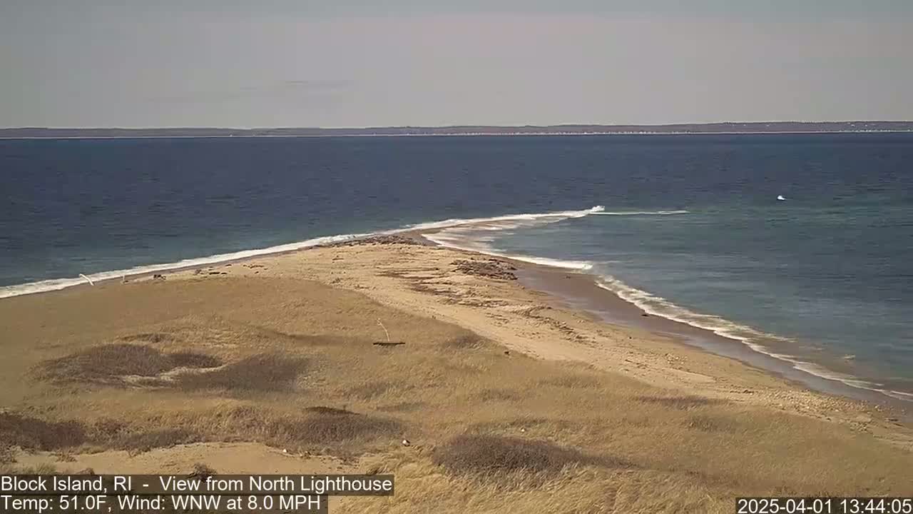 A sandy, dune-covered spit of land juts into a calm, dark-blue ocean under a clear sky.