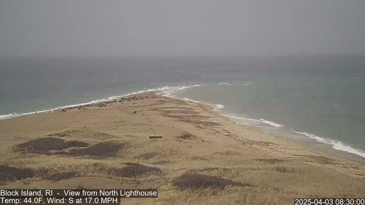 A sandy, dune-covered point of land juts into a gray, wavy ocean under an overcast sky.
