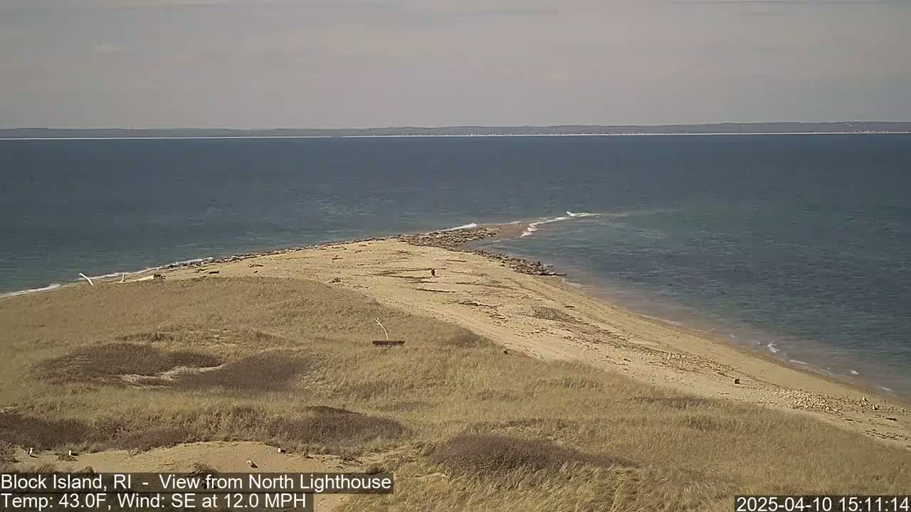 A sandy point of land juts into a calm, blue ocean under a mostly clear sky.