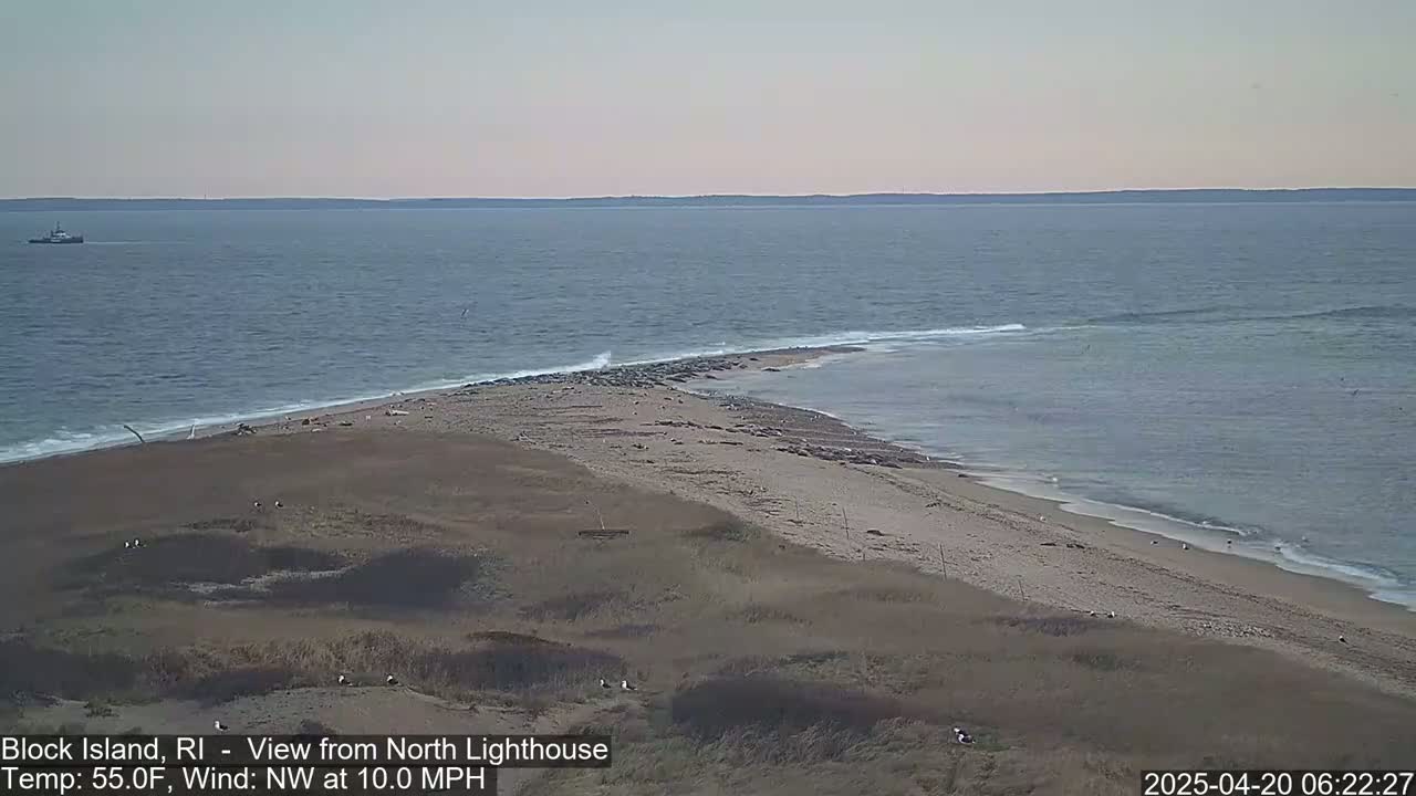 A sandy beach with a few birds and a distant boat is visible, with a calm ocean under a mostly clear sky and a northwest wind blowing at 10 mph.
