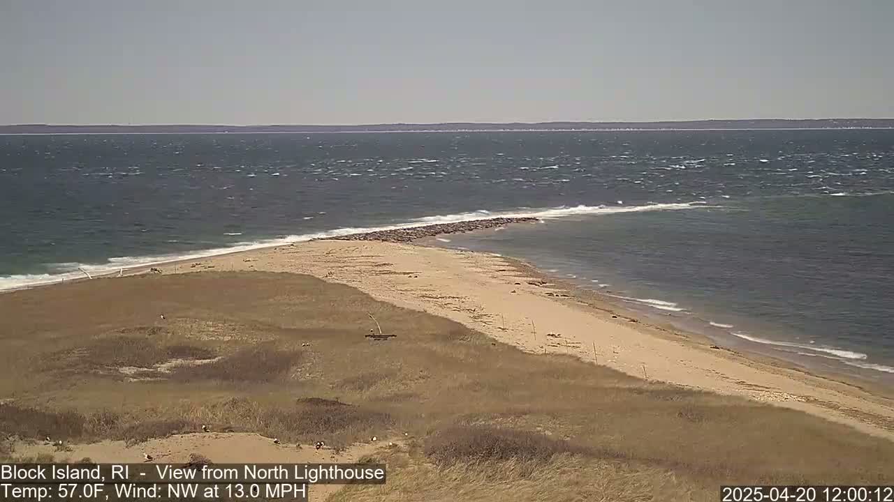 A sandy beach with dunes and a rocky jetty extends into a choppy ocean under a mostly clear sky.