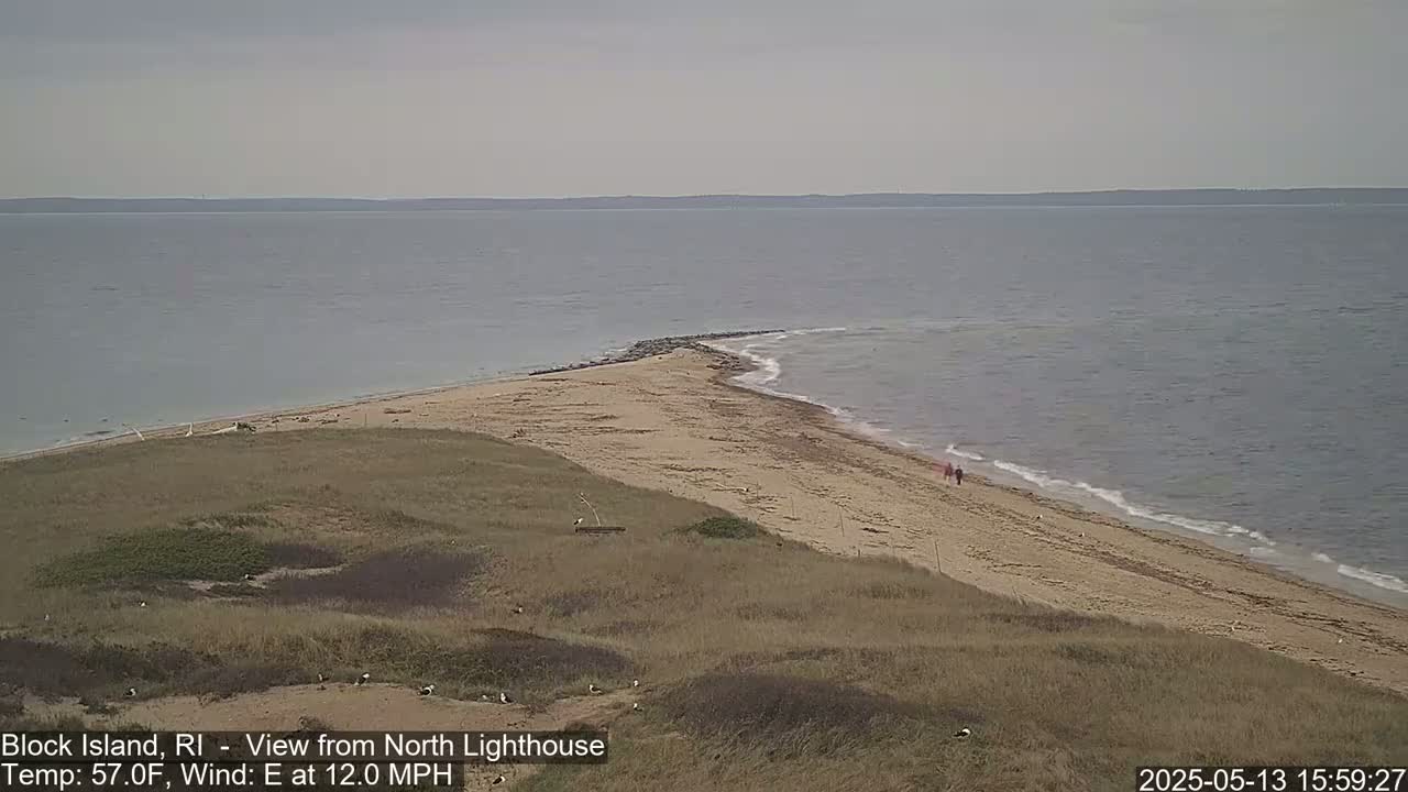 A sandy beach curves around a point of land jutting into a calm sea under a cloudy sky, with a few people visible on the beach.