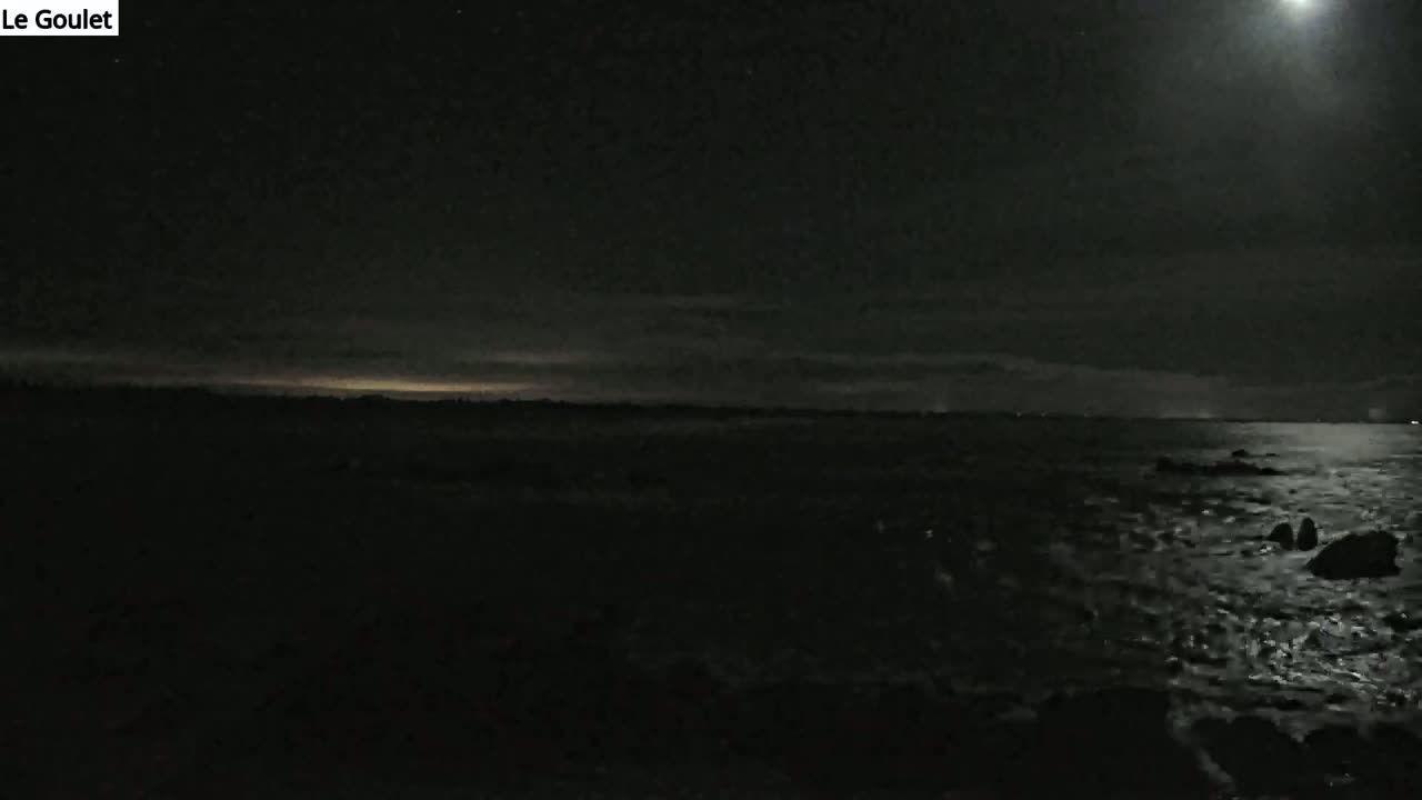 A dark, moonlit night scene shows a body of water with some rocks in the foreground and a faint horizon line in the distance.