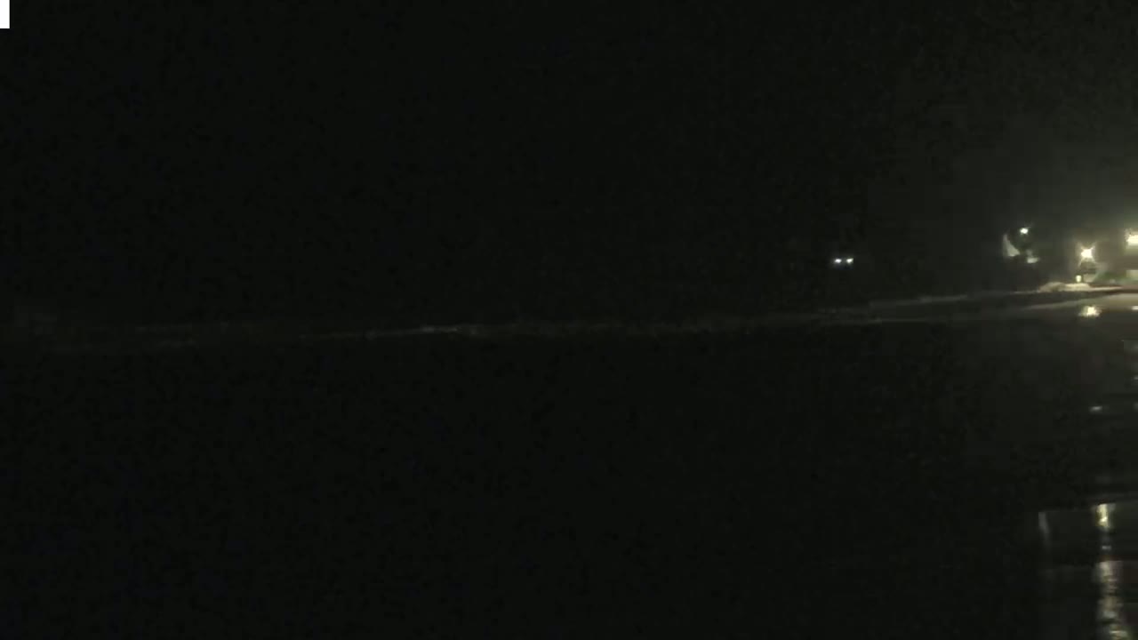 At night, a dark body of water with a faint shoreline is visible, with some dimly lit structures in the distance.