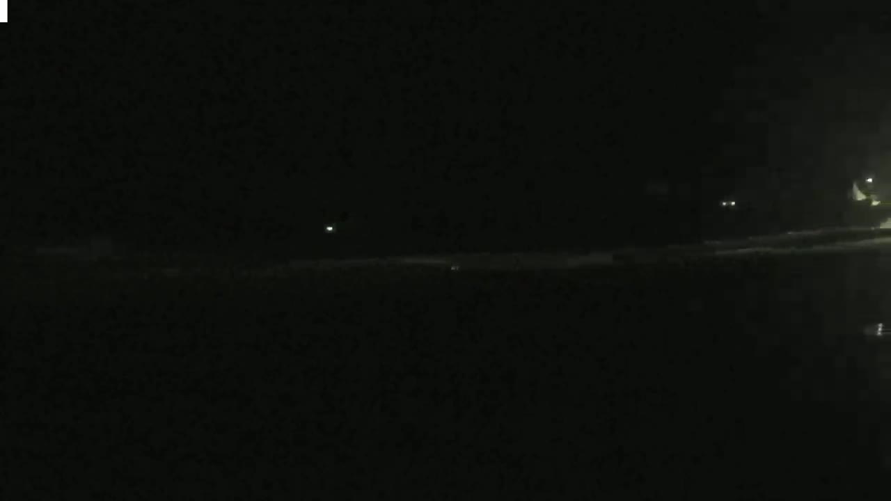 A dark, nighttime scene shows a dimly lit shoreline with faint lights in the distance.
