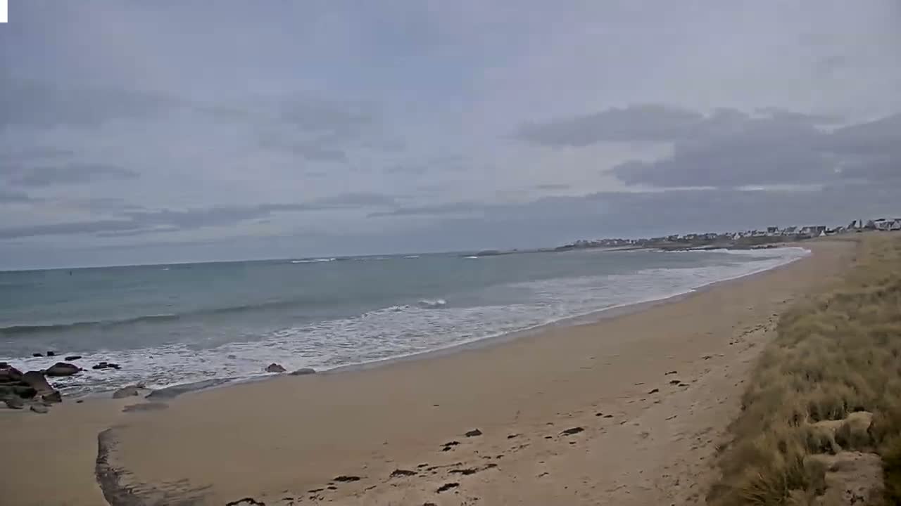 A sandy beach with gentle waves under an overcast sky stretches to a distant shoreline with buildings.