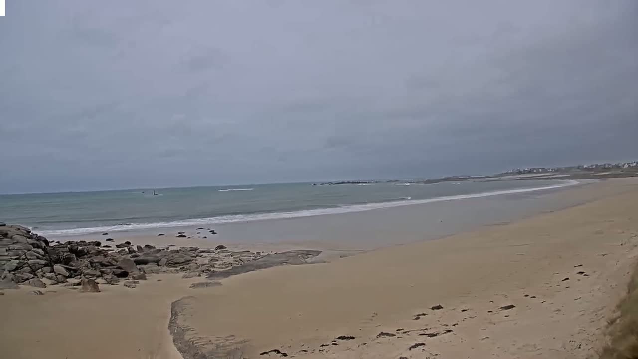 A sandy beach with dark rocks at the left edge stretches to a calm, light-green ocean under an overcast sky.