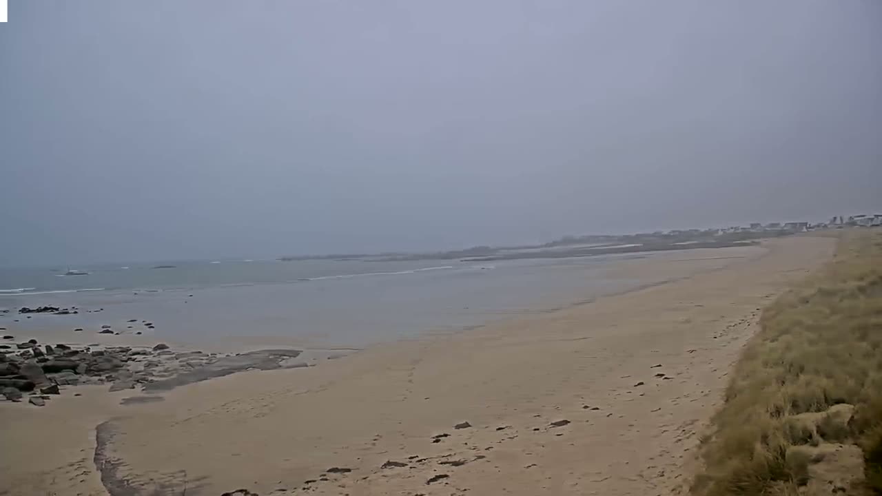 A sandy beach stretches towards a calm, hazy ocean under an overcast sky.