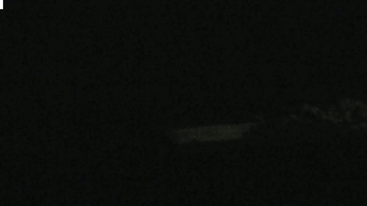A dimly lit, dark scene shows a faint, horizontal light source near the bottom, possibly from a vehicle's headlights, against a mostly dark background with indistinct shapes suggesting a nighttime outdoor setting.