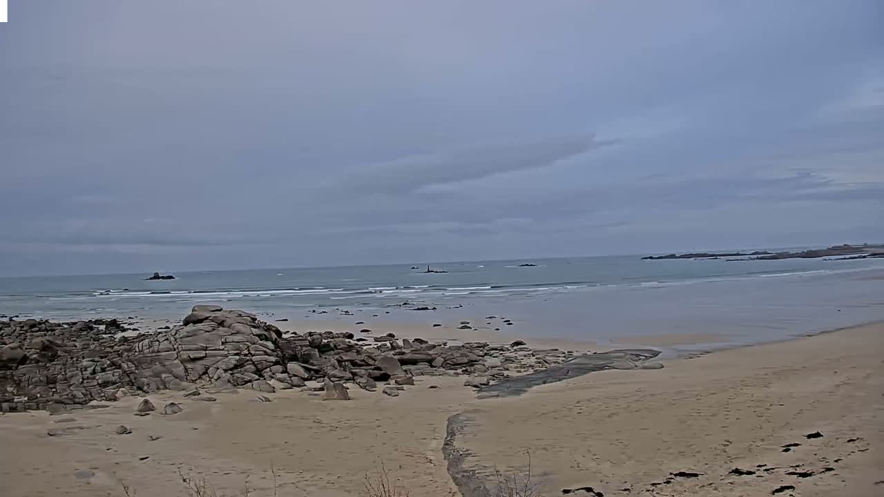 A sandy beach with dark rocks in the foreground leads to a calm ocean under an overcast sky.