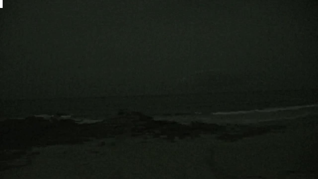 A dark, overcast sky hangs over a beach where dark, indistinct land meets a dark ocean with barely visible waves.