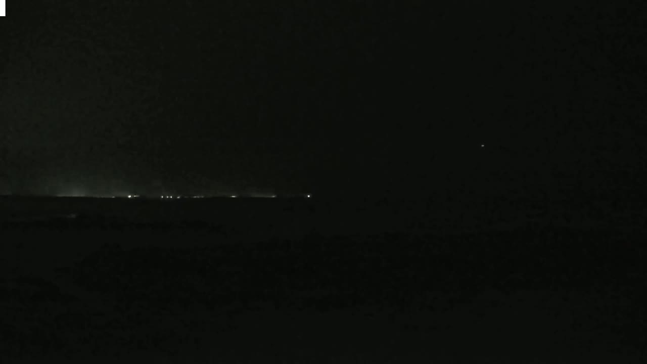 A nighttime, dark scene shows a faint line of lights on the horizon under a mostly cloudy sky.