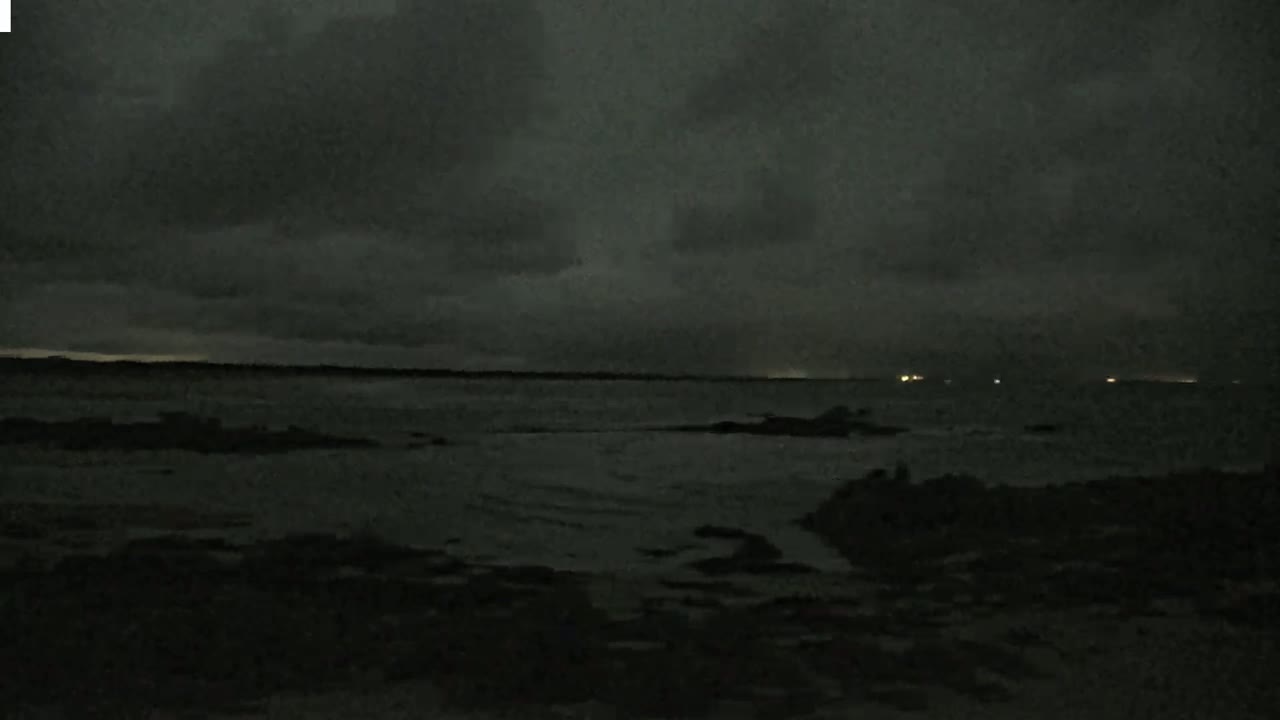 A dark, overcast night scene shows a mostly calm body of water with a dark shoreline and faint lights visible in the distance.