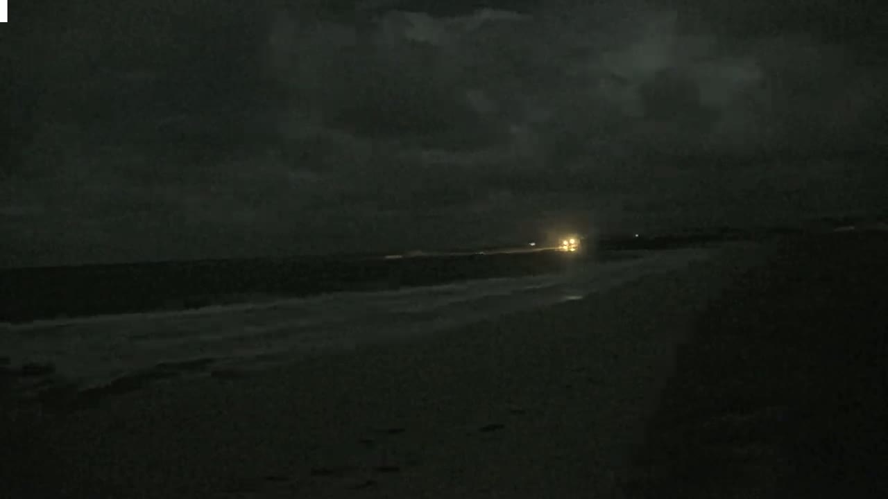 A dark, nighttime beach scene shows a dimly lit ocean with gentle waves lapping the shore under a cloudy sky.