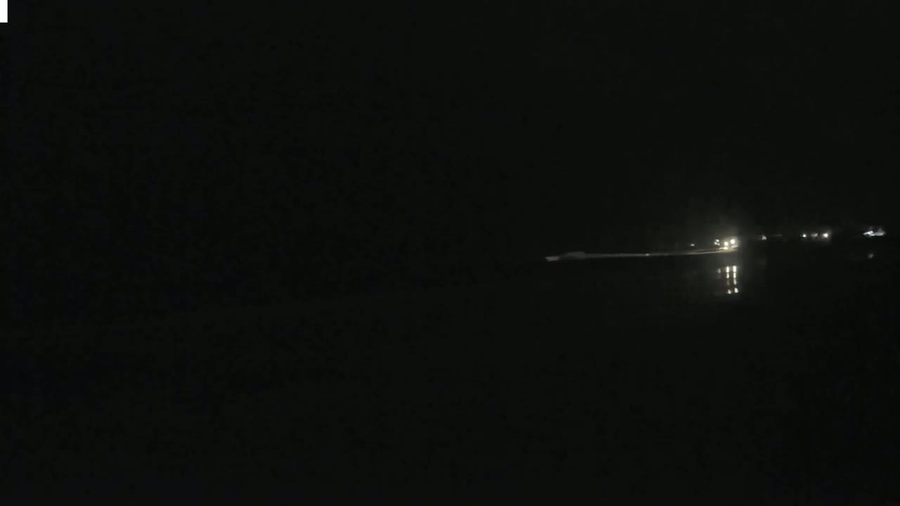 A dark, nighttime scene shows a body of water reflecting faint lights from a distant roadway.