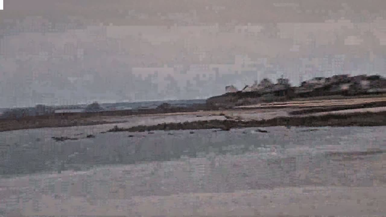 A calm, overcast day reveals a flat, sandy shoreline with a few buildings visible in the distance on a raised area.