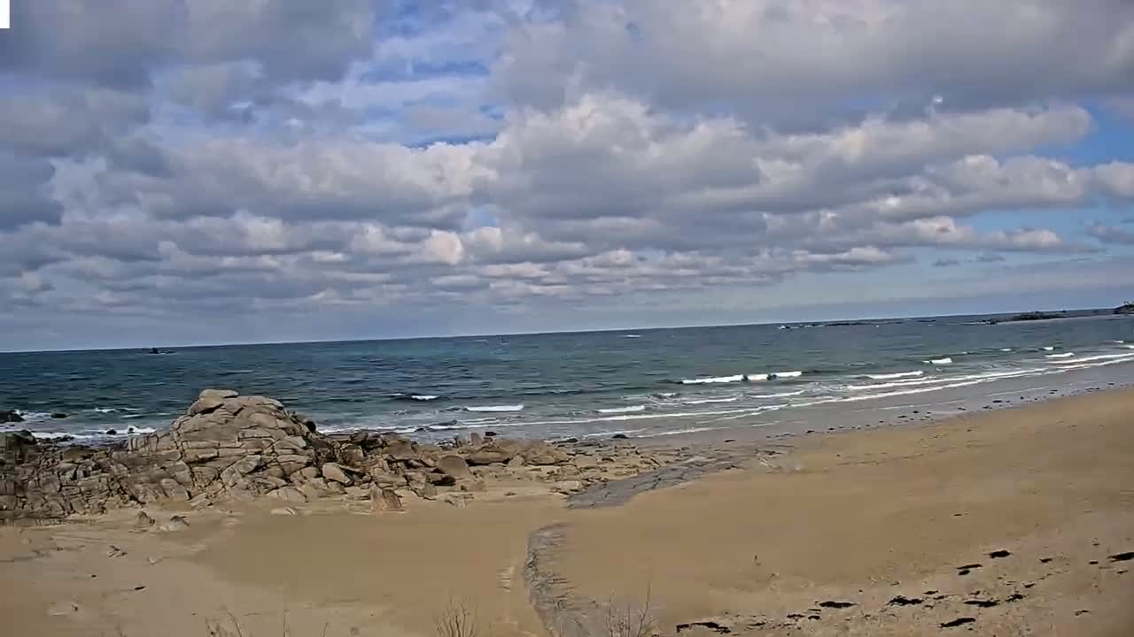A sandy beach with rocky outcrops meets a gently wavy ocean under a partly cloudy sky.