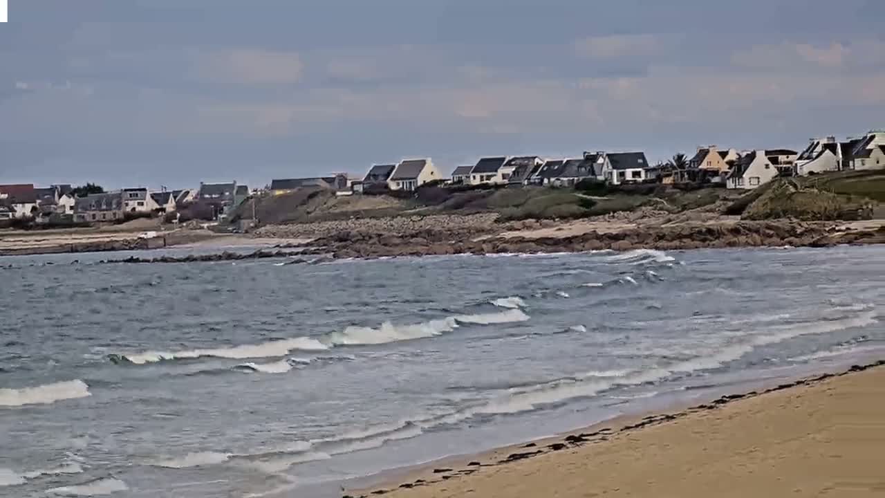 A sandy beach with small waves laps against a rocky shoreline where a cluster of houses sits on a grassy hillside under a mostly cloudy sky.