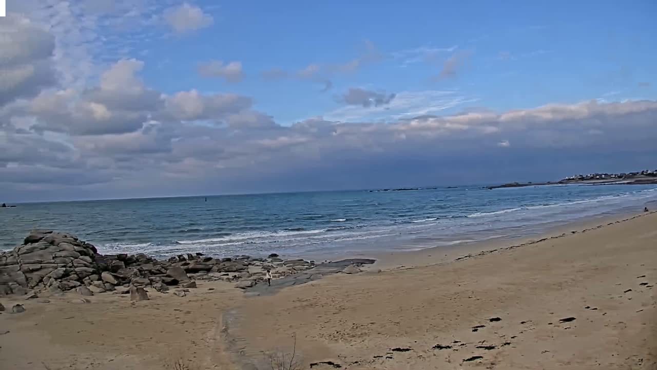 A sandy beach with rocky outcrops stretches to a calm ocean under a partly cloudy sky.