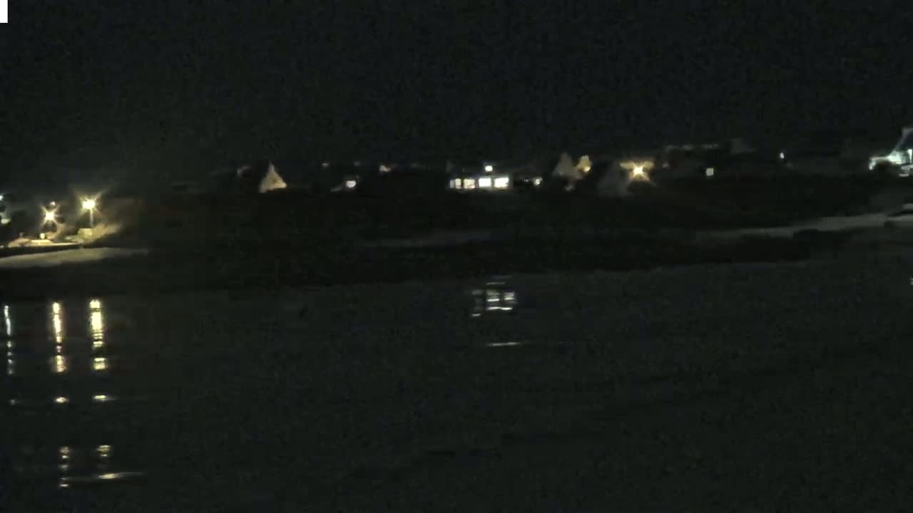 At night, several illuminated houses are visible across a dark body of water, whose surface reflects the lights.