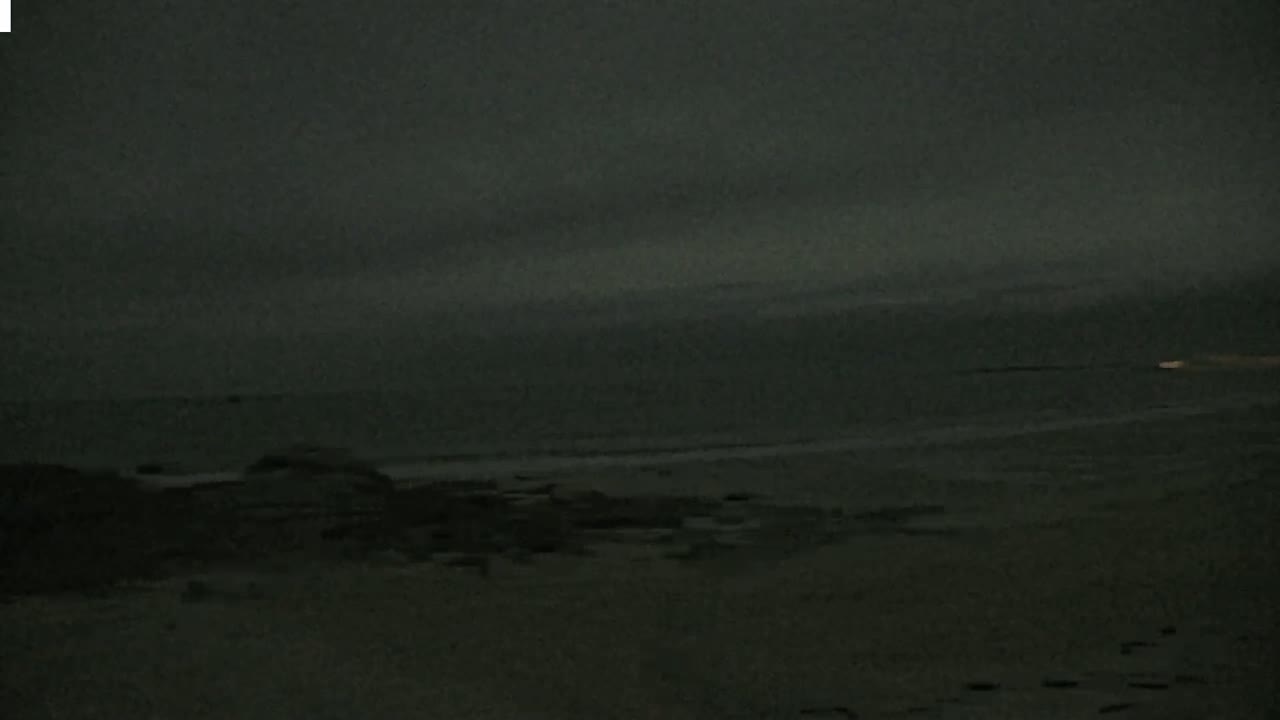 A dark, overcast sky hangs over a dimly lit beach with dark rocks and sand.