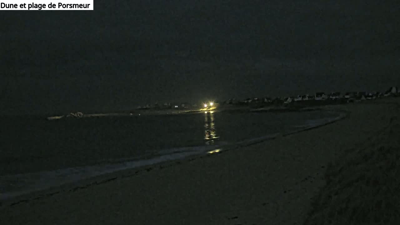 A dark, nighttime beach scene shows a gently lapping ocean and a dimly lit town in the distance under a cloudy sky.