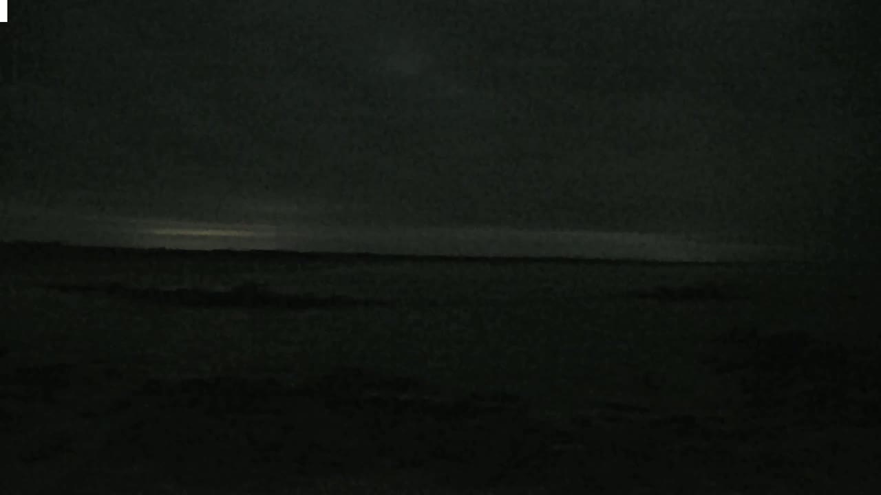A dark, nighttime scene shows a body of water under a mostly cloudy sky with a faint, light horizon.