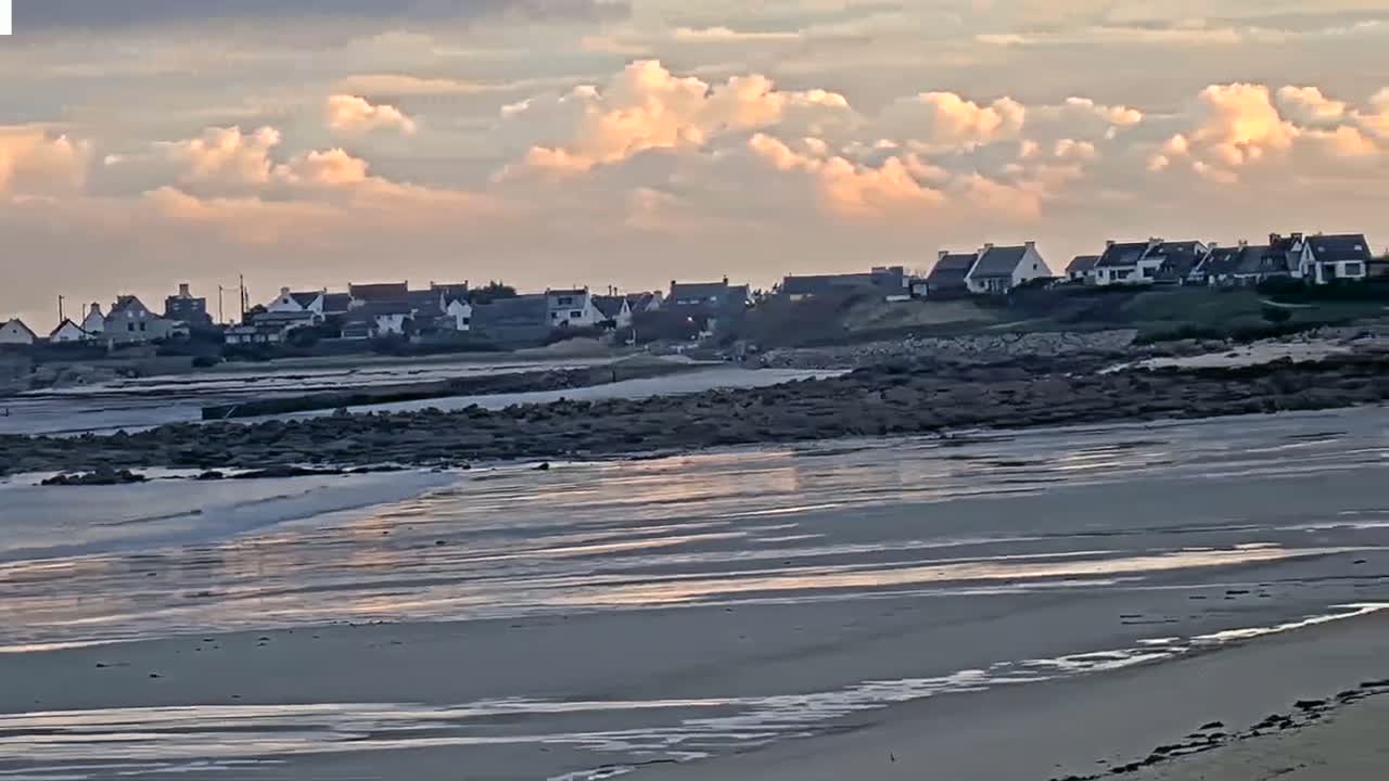 A coastal village sits atop a rocky shoreline under a partly cloudy, sunset sky, with a wide, sandy beach extending into the calm ocean.