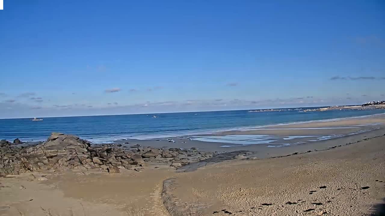 A sandy beach with dark rocks in the foreground stretches to a calm, light blue ocean under a clear blue sky with a few clouds.