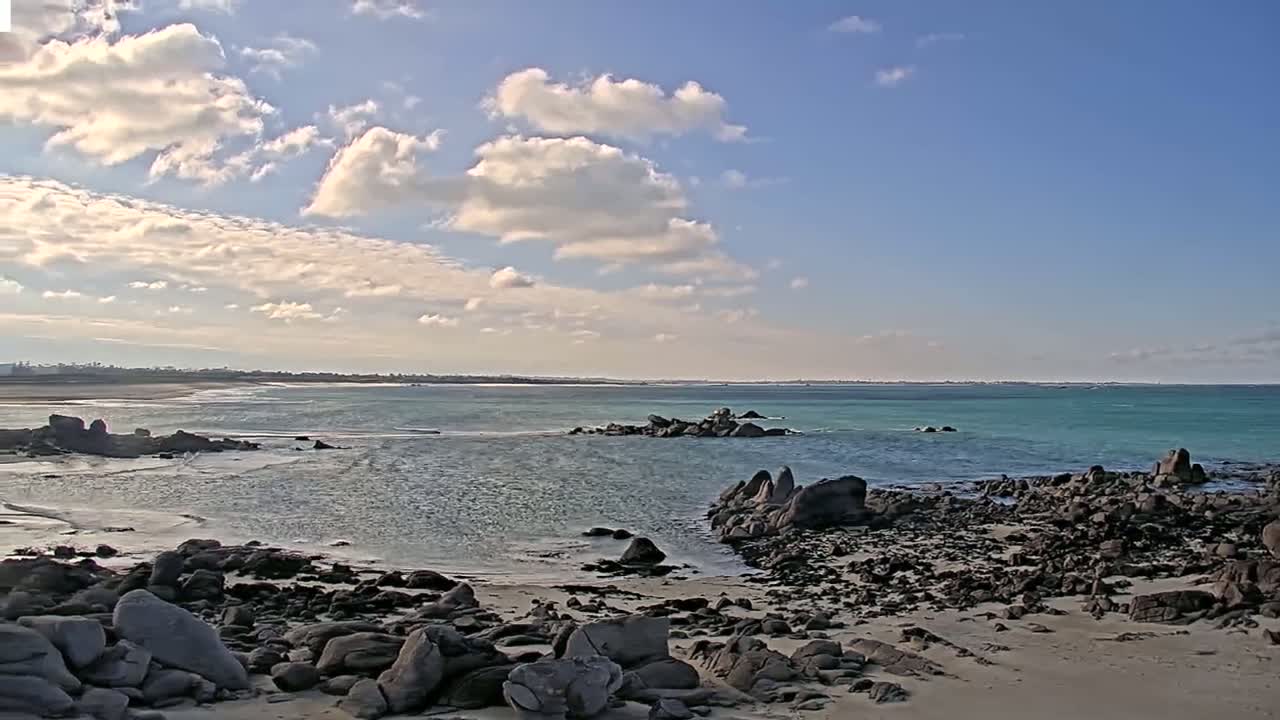 A rocky shoreline meets a calm, teal ocean under a partly cloudy sky.