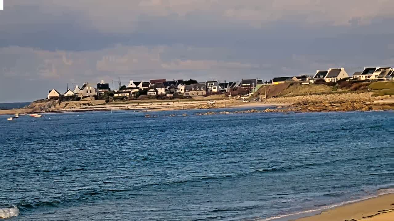 A coastal village with houses on a rocky shoreline is visible across a calm, blue ocean with small waves lapping a sandy beach in the foreground under a mostly cloudy sky.