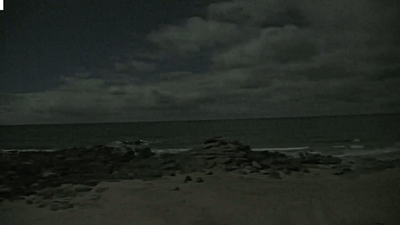 A dark, cloudy night scene shows a rocky shoreline meeting a calm, dark ocean.