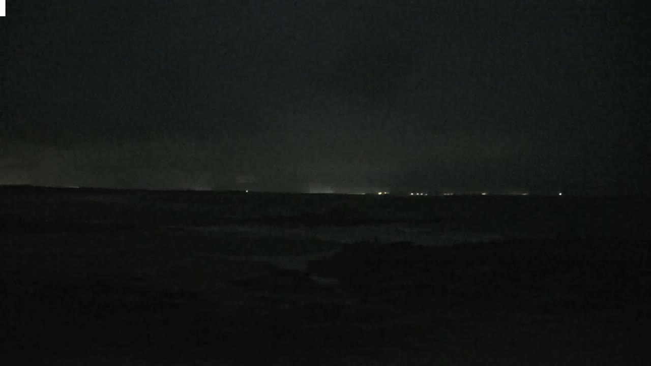 A dark, nighttime ocean scene shows a faint line of lights on the horizon under a cloudy, dark sky with slightly agitated waves.