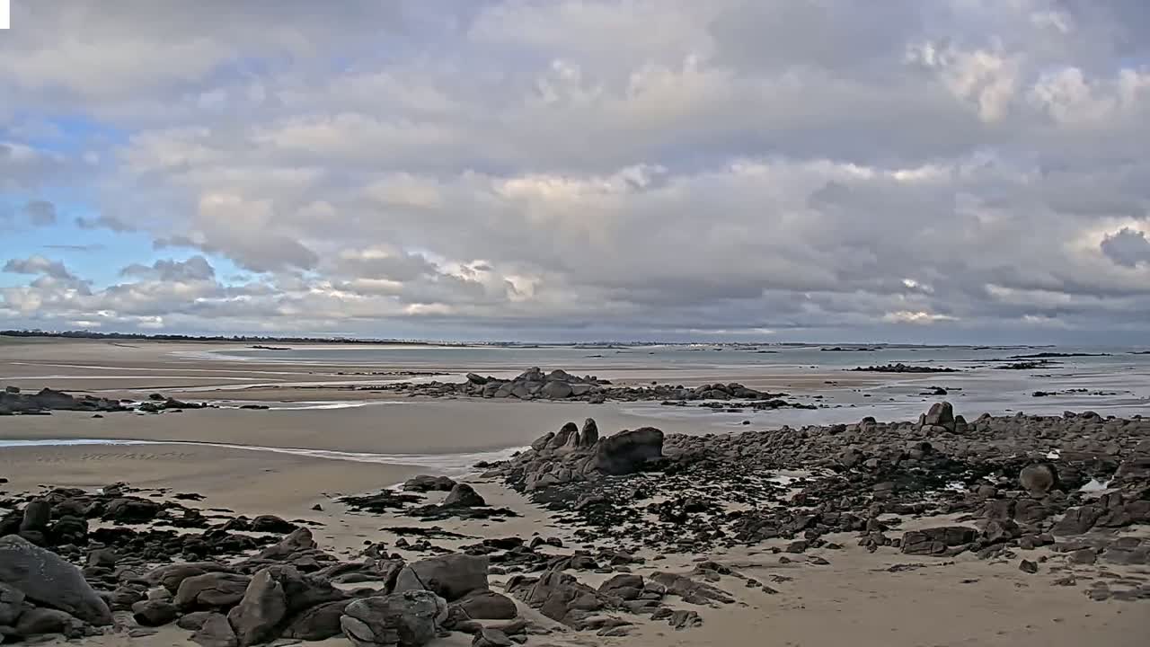 A sandy beach with dark rocks scattered across it stretches towards a calm ocean under a cloudy sky.