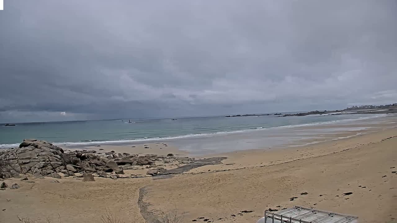 A sandy beach with rocky outcrops stretches to a calm, light-green ocean under a cloudy sky.