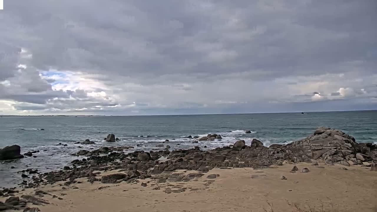 A sandy beach with dark rocks extends into a moderately wavy ocean under a mostly cloudy sky.
