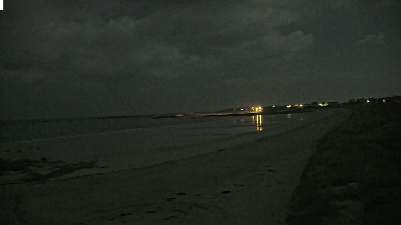 A dark, sandy beach at night under a cloudy sky is dimly lit by distant lights.