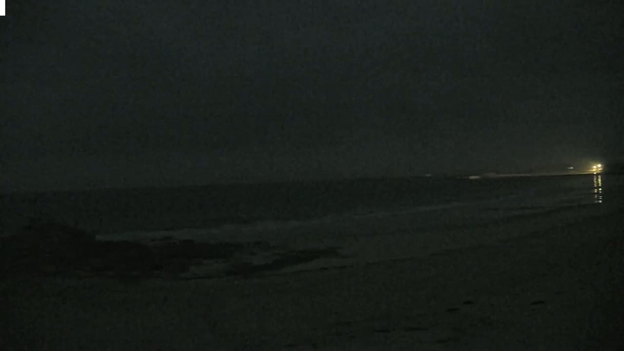 A dark, nighttime beach scene shows a calm ocean with a distant light source and gently lapping waves.