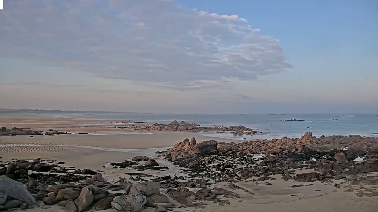 A sandy beach with dark rocks and seaweed stretches to a calm ocean under a partly cloudy sky.