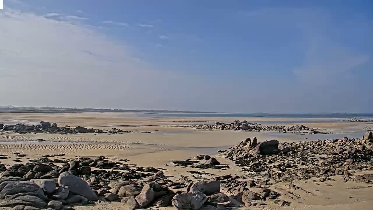 A sandy beach with dark rocks and shallow water stretches to a calm ocean under a mostly clear blue sky.