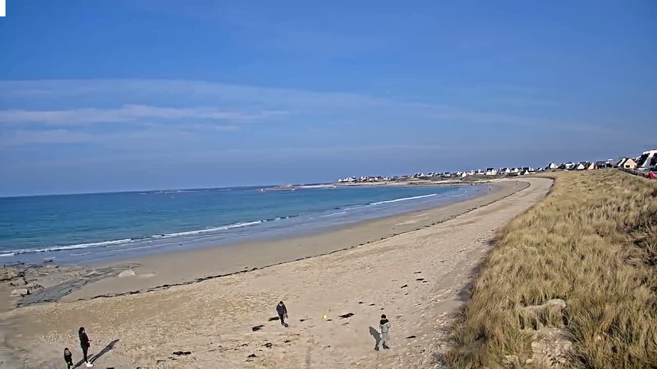A long sandy beach curves gently to the left, meeting a calm, light blue ocean under a mostly clear, bright blue sky; a few people are seen on the beach.