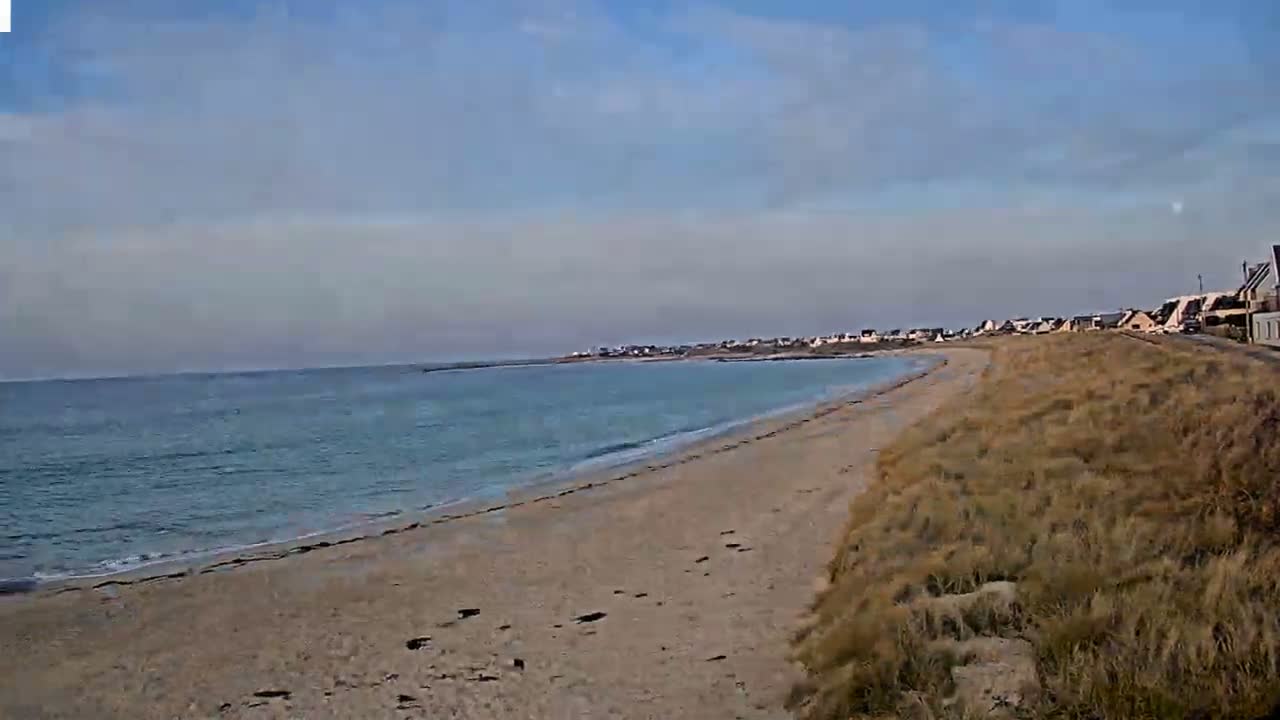 A sandy beach stretches alongside a calm, light-blue ocean under a mostly clear sky, with a small town visible in the distance.