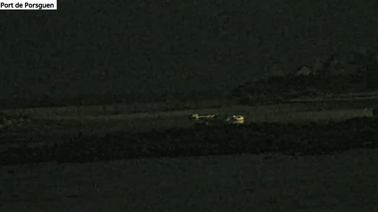 At night, a dark body of water reflects a faint, yellowish glow from a distant light source, with dark land visible in the foreground and background.