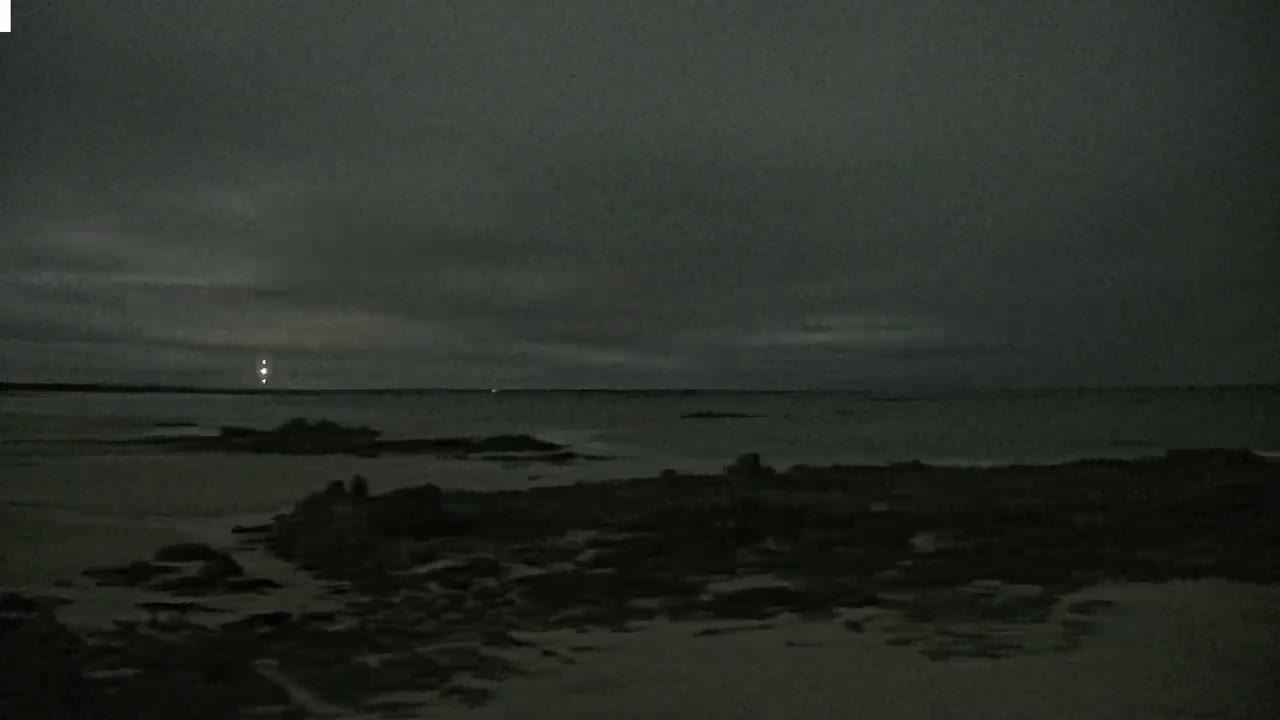 A dark, overcast night scene shows a mostly calm body of water with a dark shoreline and a faint light visible in the distance.